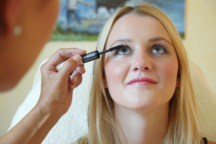 Eine blonde Frau mit hellem Teint lächelt sanft, während ihr Wimperntusche aufgetragen wird
